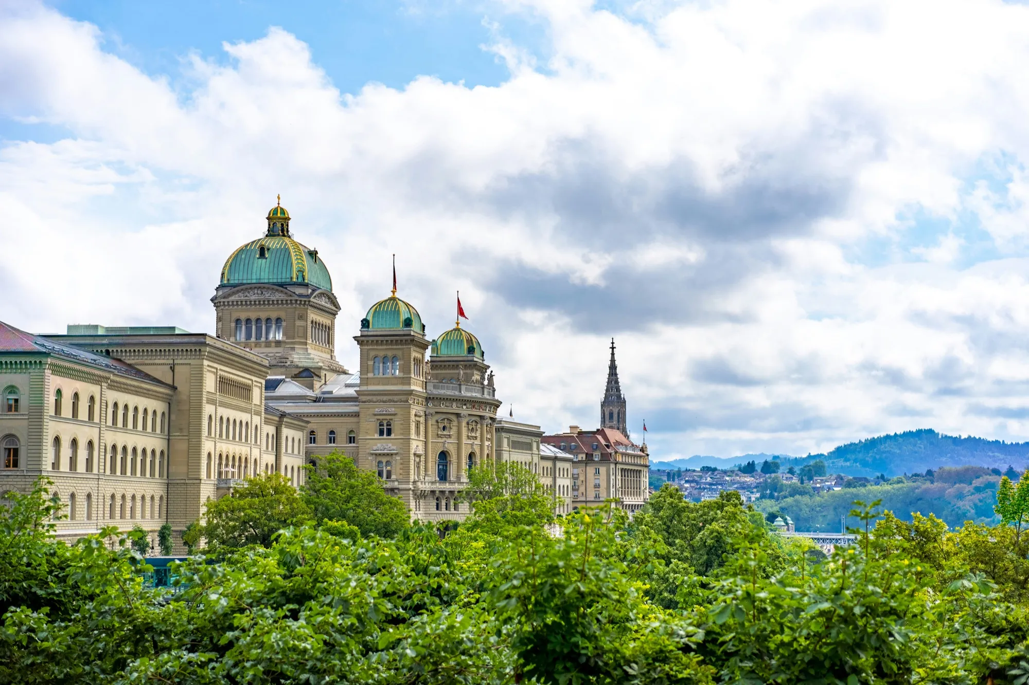 Das Bundeshaus in Bern mit grünen Kuppeln über Bäumen, blauer Himmel mit Wolken