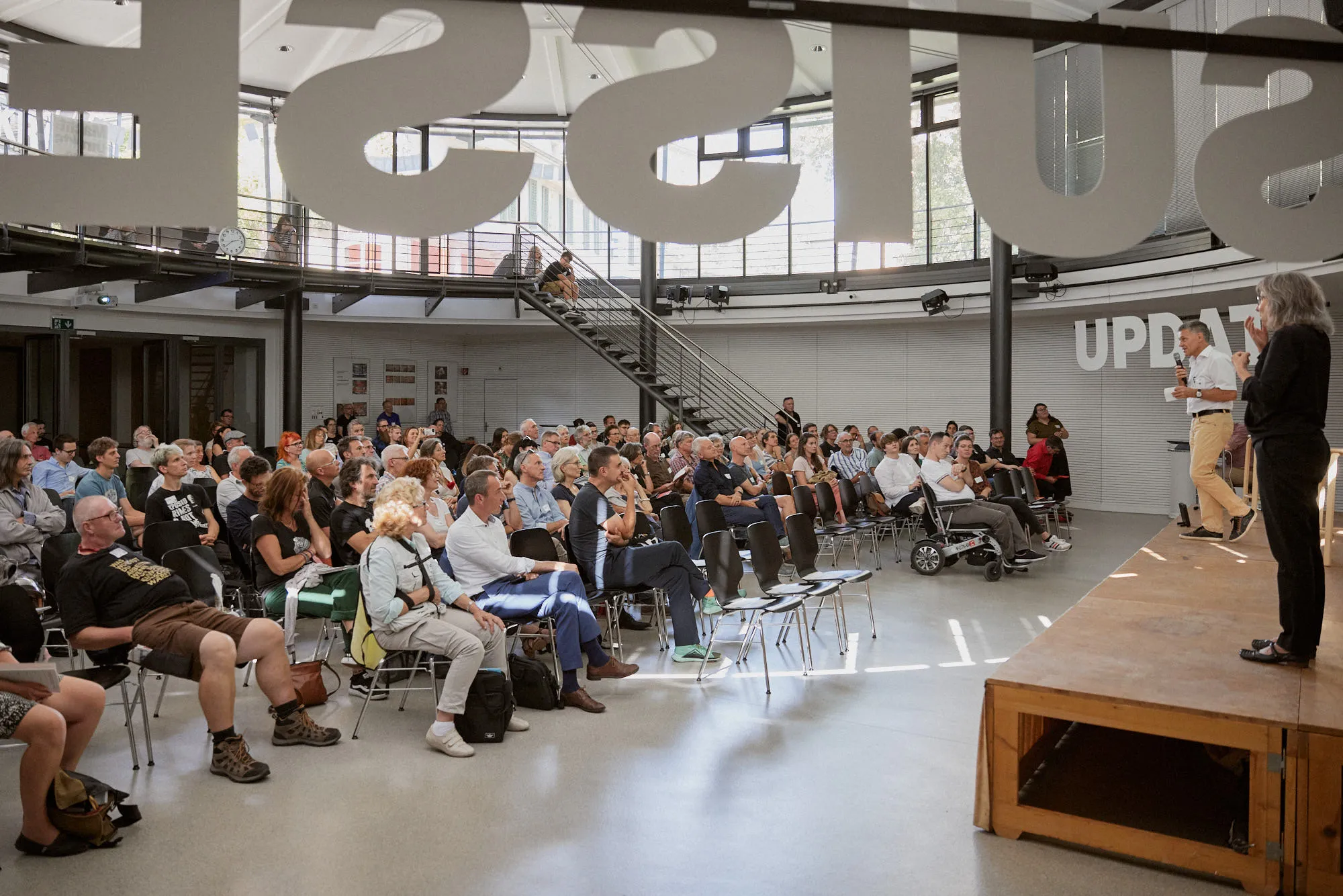 Volle Halle mit Publikum, vorne eine Bühne mit dem Schriftzug «UPDATE», an der Decke grosse Buchstaben